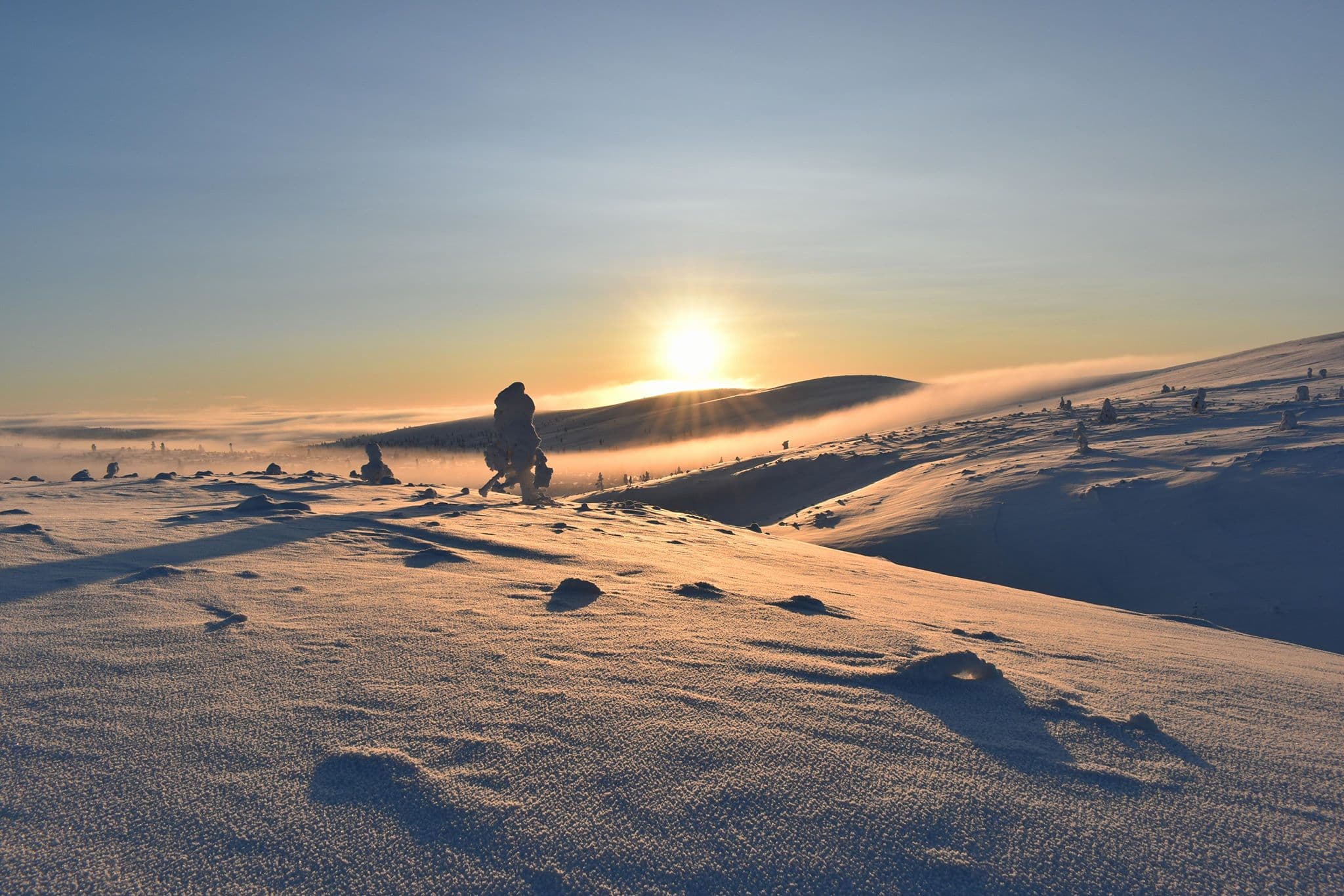 Bild: Tero Turpeinen Solen skiner bakom den snötäckta fjällen. På fjällens sluttningar finns bara några snötäckta tallar.