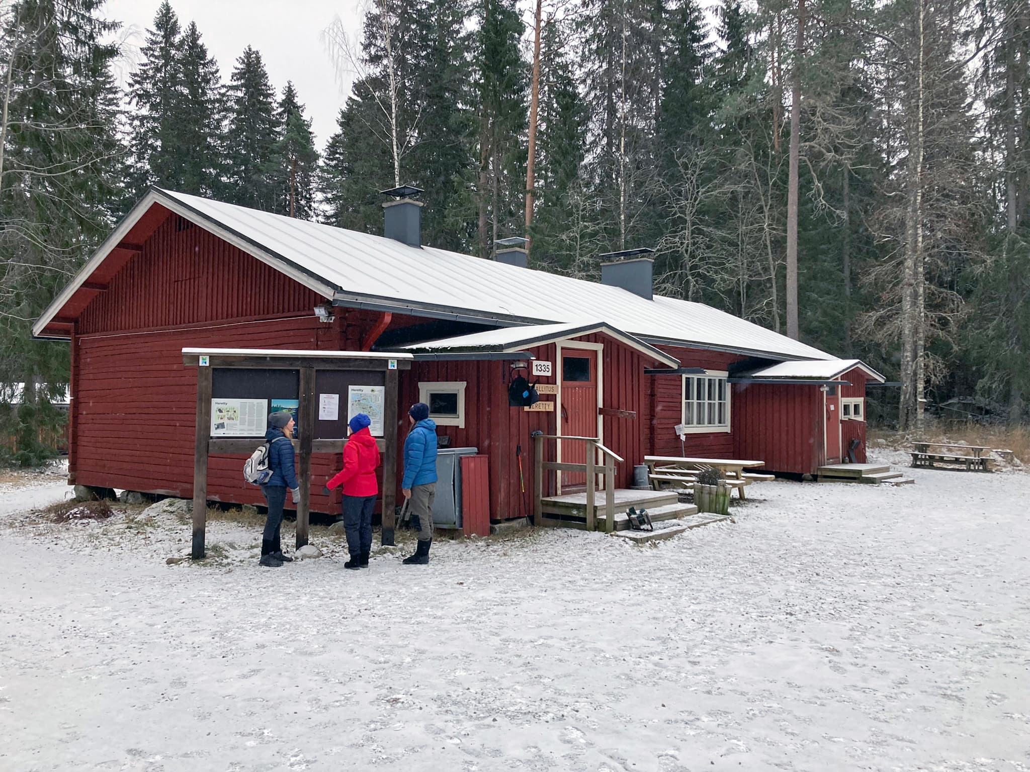 Kuva: Maija Mikkola Kolme retkeilijää tutkii opastetaulua punaisen puurakennuksen vieressä.