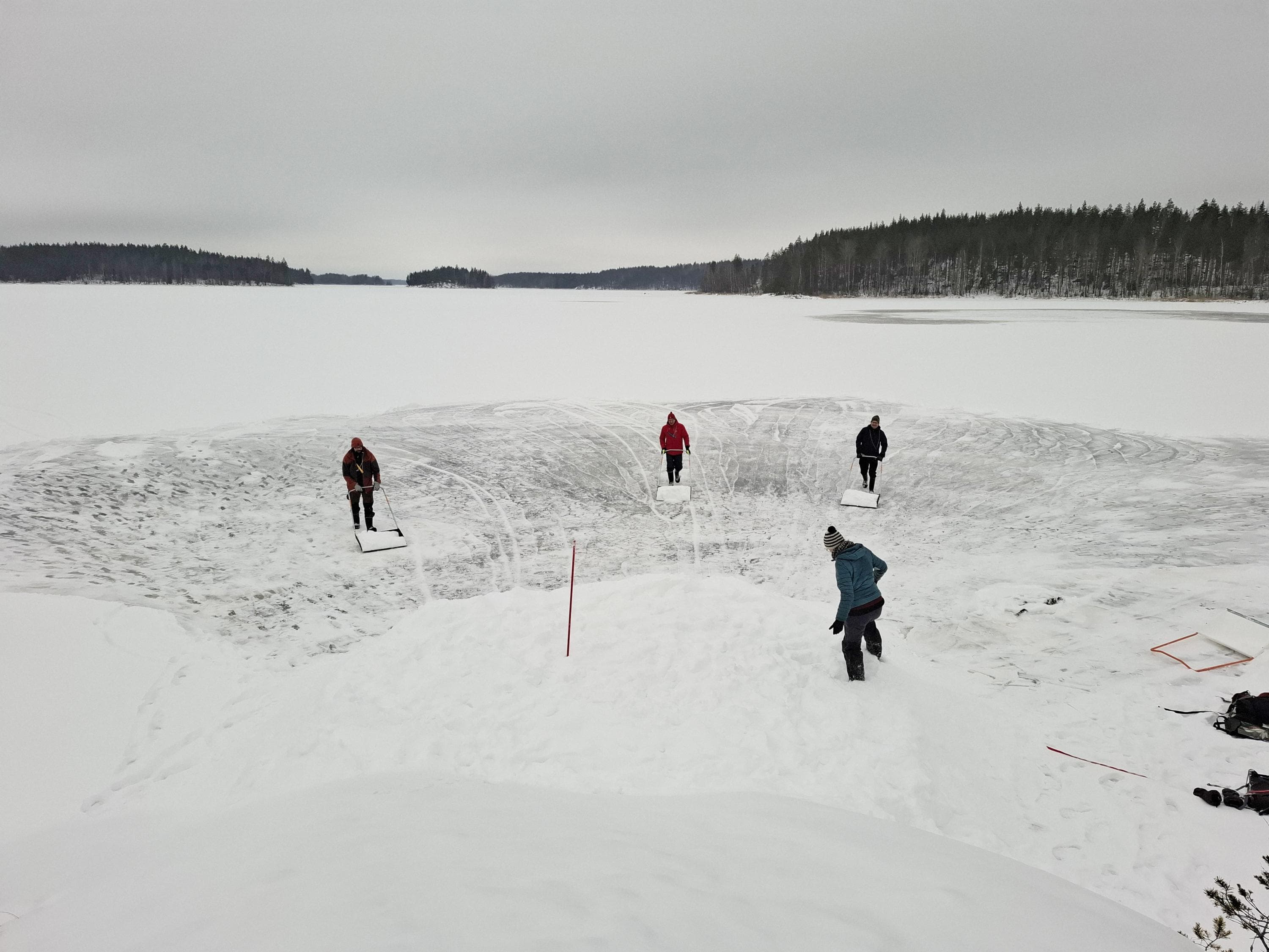 Kuva: Riikka Alakoski Kolme kolaajaa tuomassa järven jäällä lunta kohti kinosta, jonka päällä seisoo yksi ihminen.