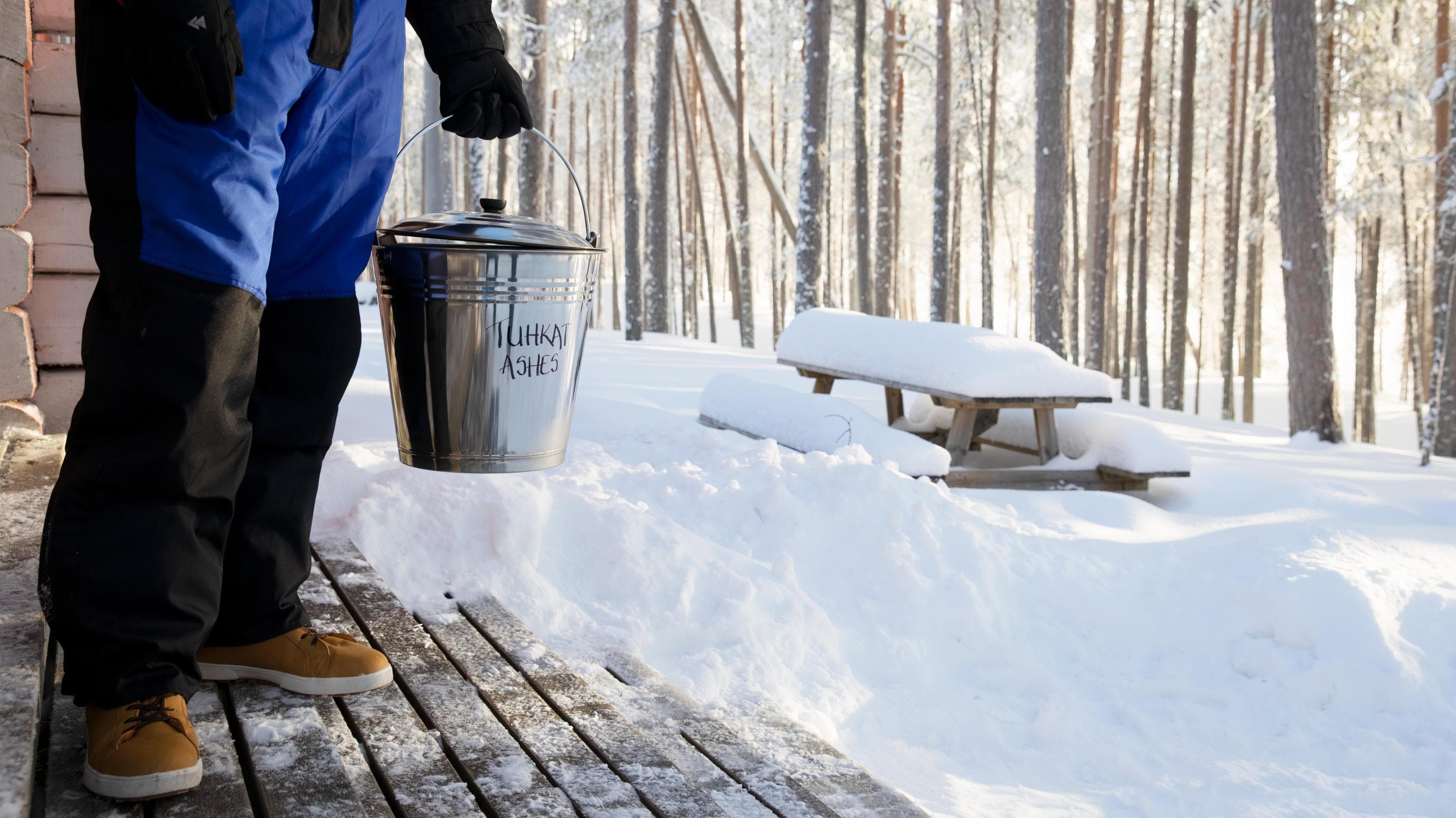 Kuva: Katri Lehtola, Kuvatoimisto Keksi Henkilö seisoo tuvan etuterassilla tuhkasanko kädessä. Aurinko paistaa ja on talvi.
