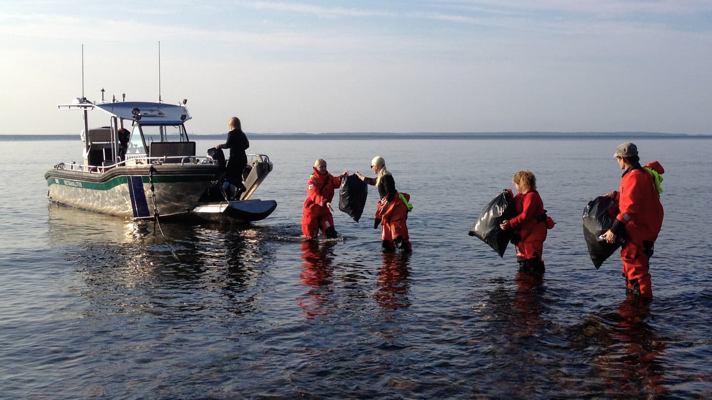 Image: Maiju Lanki Bags of trash are being loaded into a boat by a human chain. The boat is standing a short distance from the shore. The water is calm.