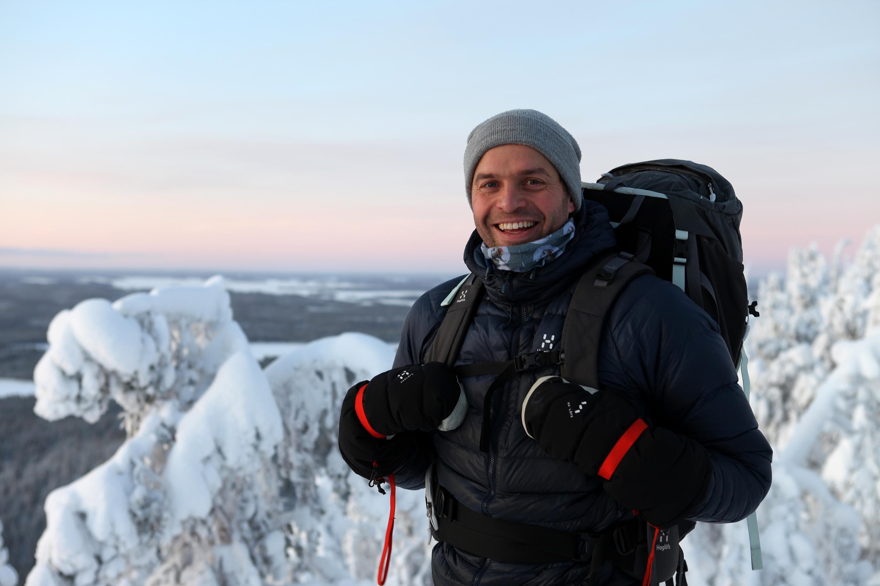 Retkeilijä katsoo kohti, hänellä on selässään reppu ja hän hymyilee. On talvi.