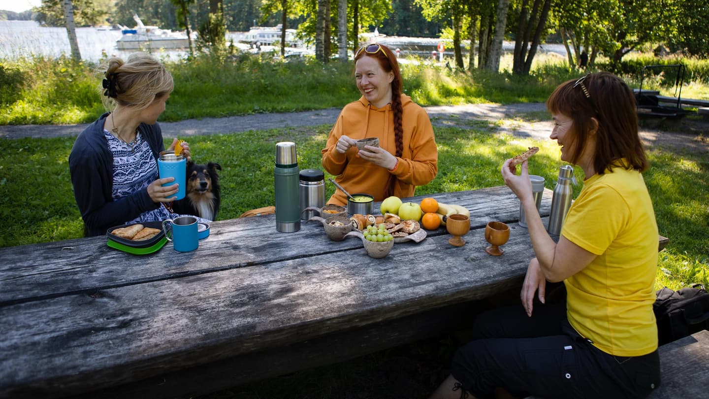 Image: Ulla Keituri Three cheerful hikers are sitting at a wooden picnic table having a snack break. A dog is with them as well.