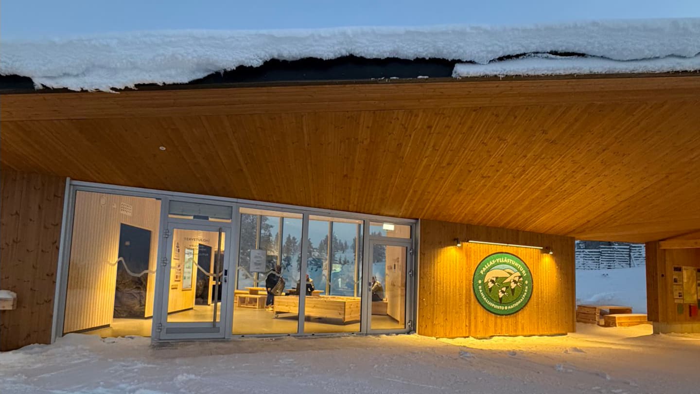 Image: Heta Kylmämaa A single-storey wooden building with the Pallas–Yllästunturi emblem on the wall. There is also an opening in the wall, and on the adjacent wall various information panels are mounted. Through the large glass windows you can see into the illuminated space where people are present.