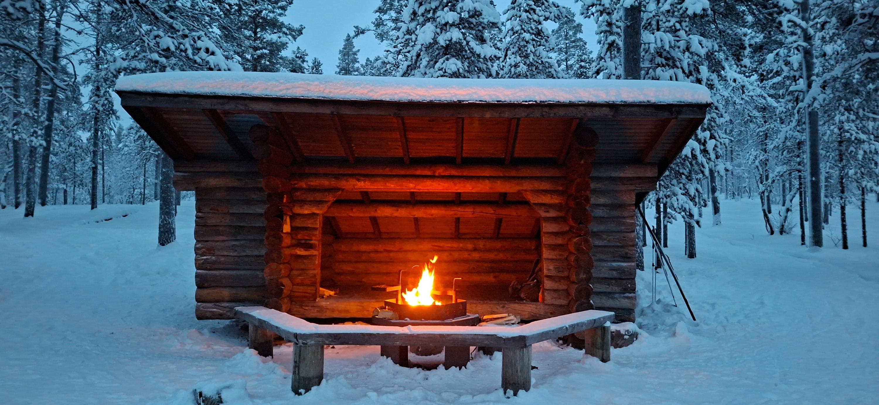 Kuva: Maria Makkonen Laavun edessä palaa tuli. Ympärillä lumisia mäntyjä. Maassa on lunta.