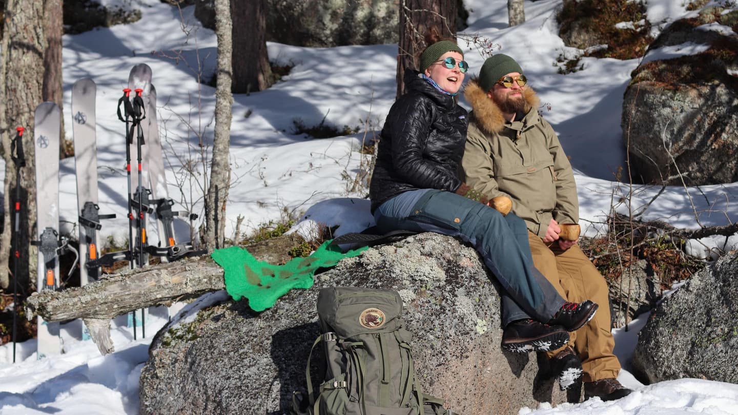 Image: Tommi Lehtinen Two hikers are sitting on a large rock in the spring sunshine. There is still snow on the ground, and the hikers are moving on sliding snowshoes.