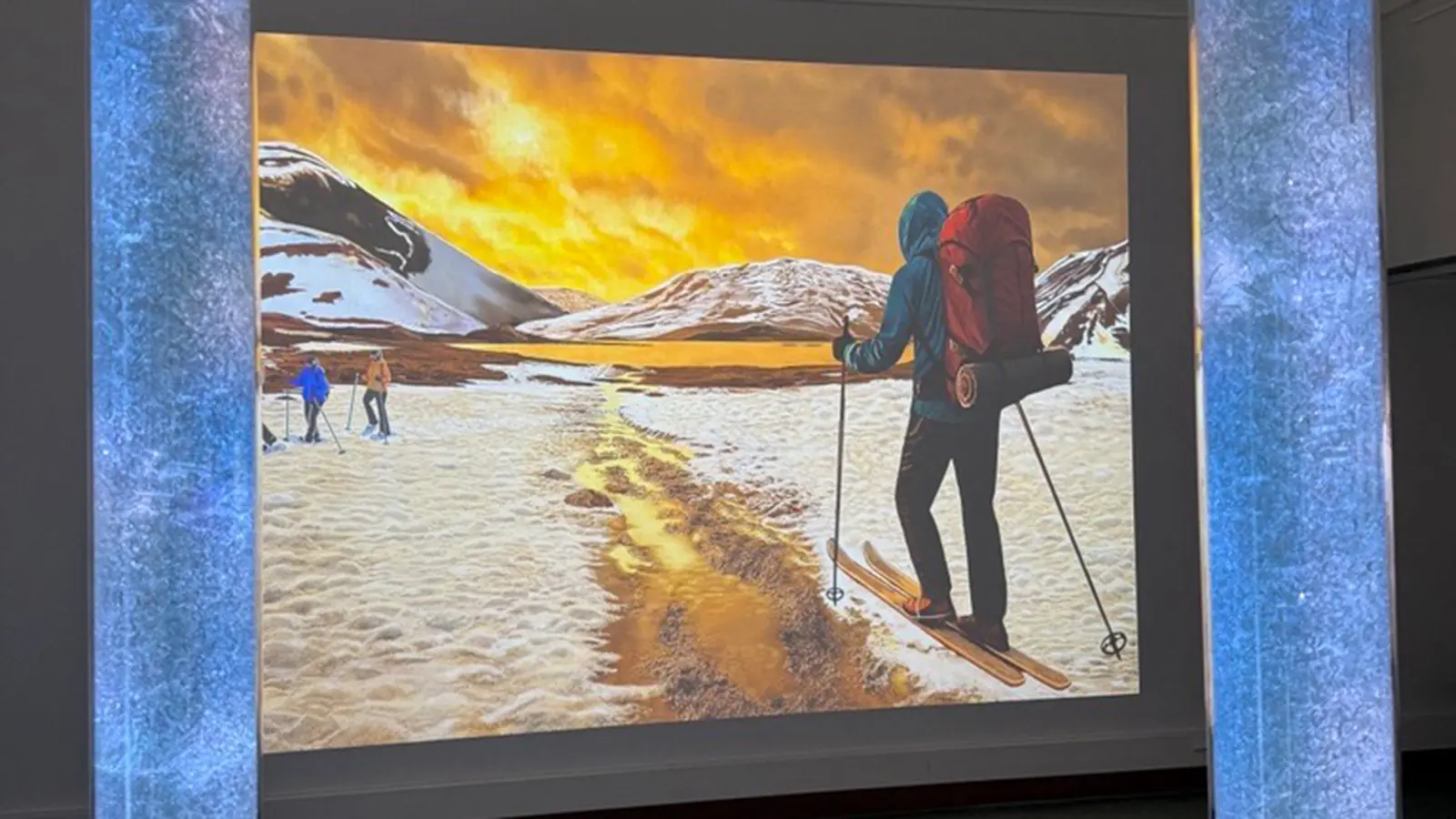Kuva: Outi Ala-Härkönen Teos näyttelyssä. Teoksessa hiihtäjiä ja osin sulanutta lunta.