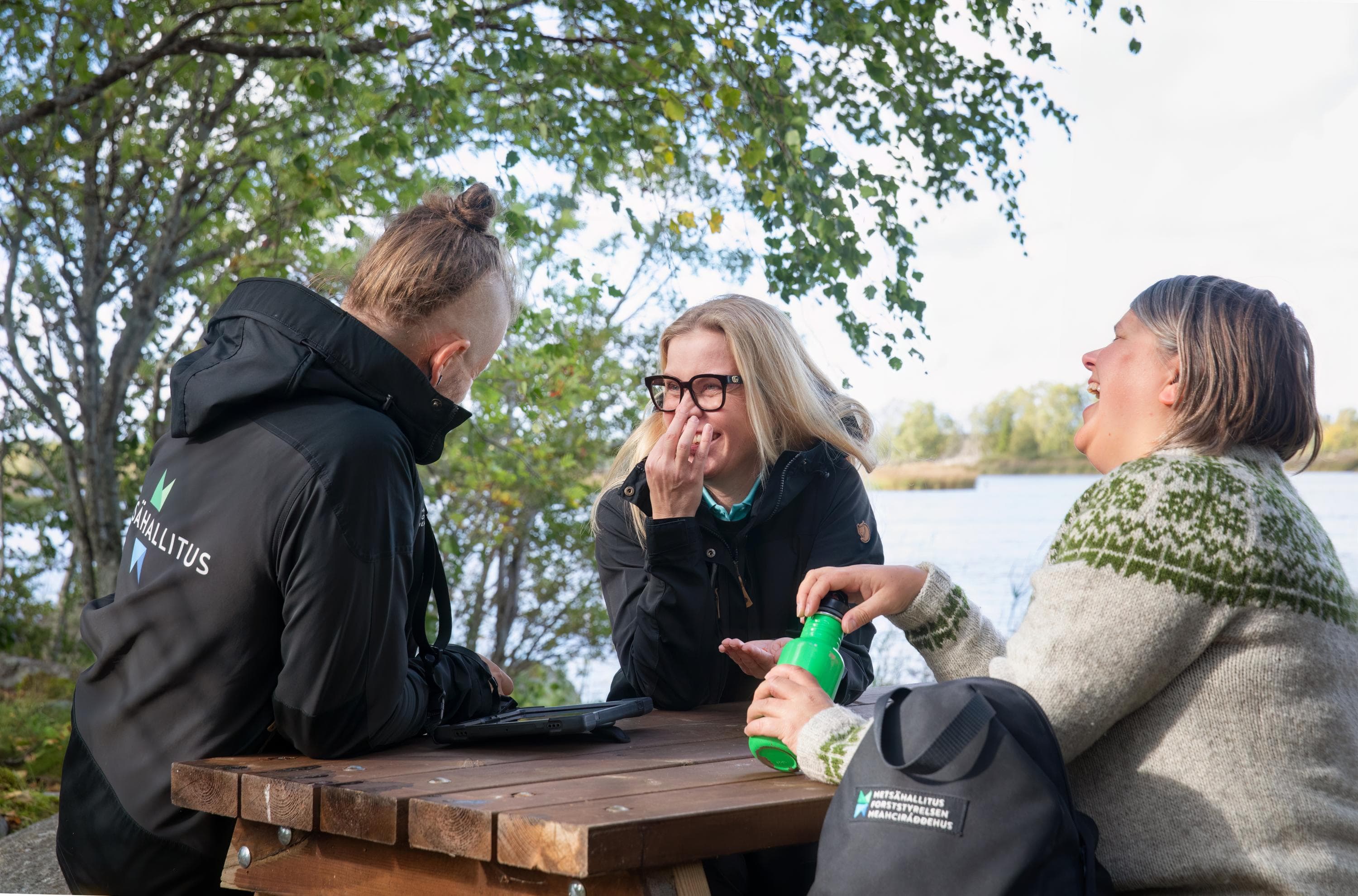 Kuva: Katri Lehtola / Keksi Kolme henkilöä istuu ulkona puisen pöydän ääressä retkivaatteissa ja juttelee nauraen.