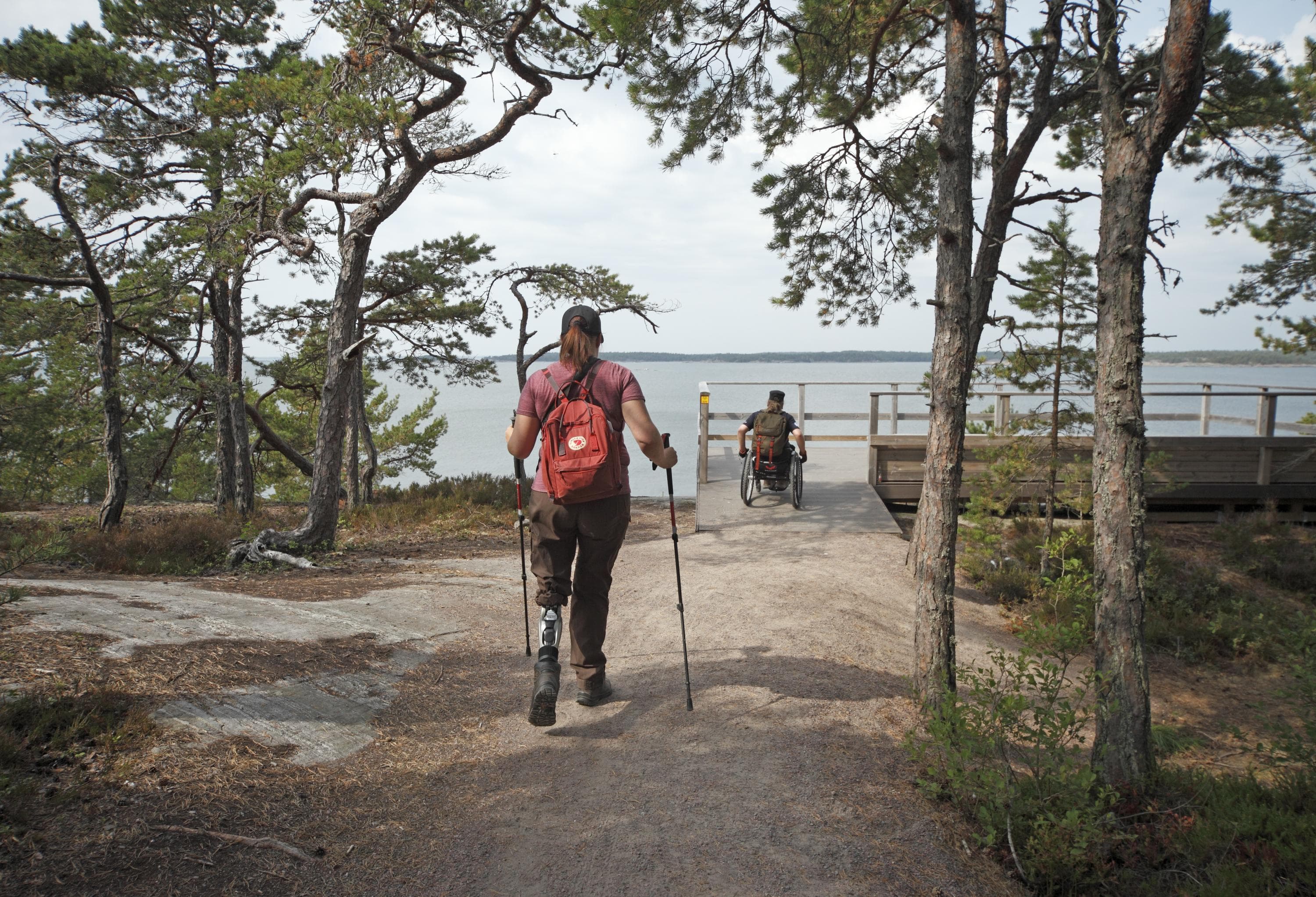 Kuva: Jussi Helimäki Kaksi retkeilijää menossa esteetöntä reittiä pitkin meren rannan esteettömälle näköalatasanteelle kesäpäivänä. Edellä kulkija kelaa pyörätuoliaan perässään patikoija sauvakävellen.