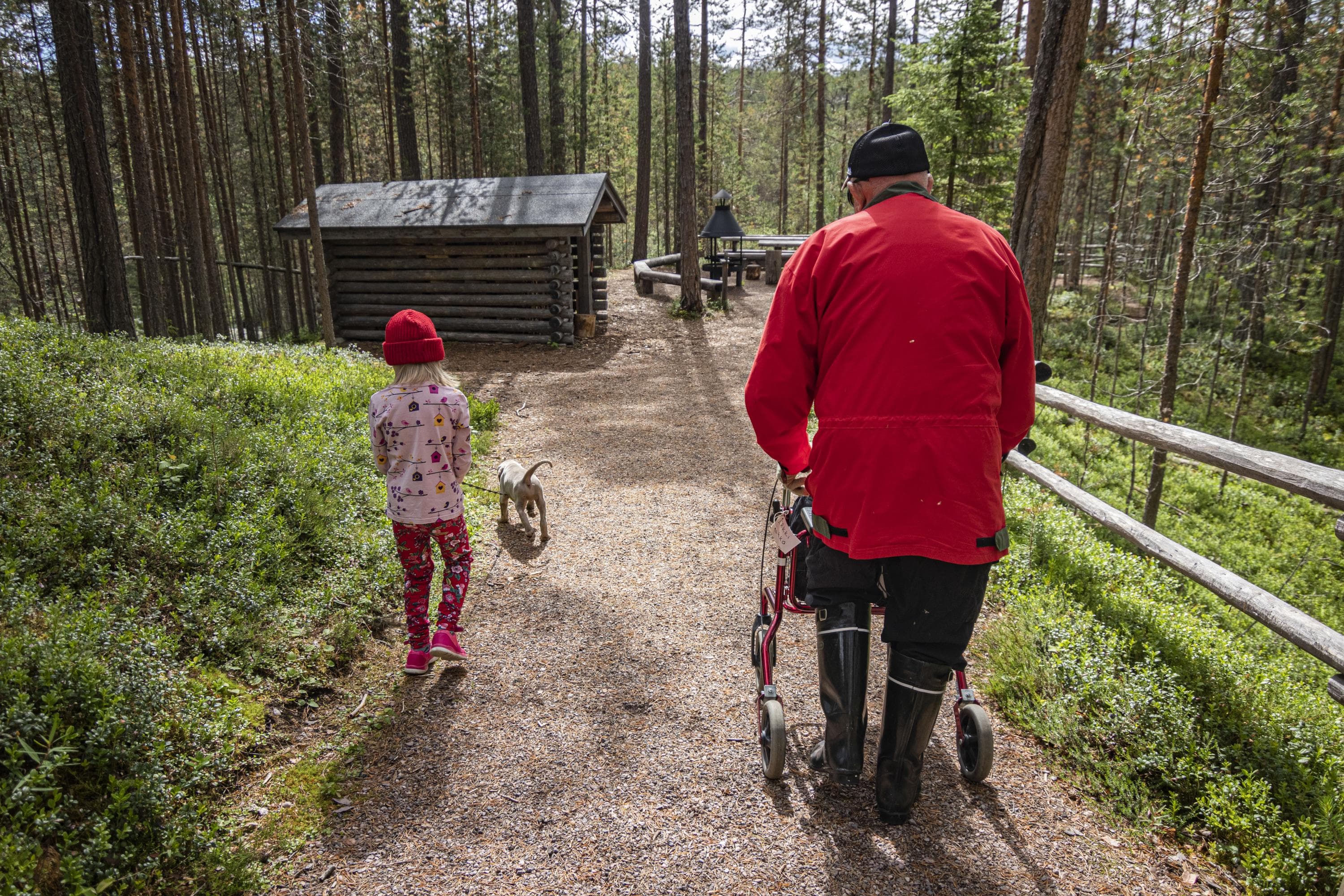 Kuva: Jonna Kalliomäki Etualalla vaeltaja rollaattorin kanssa sekä lapsi ja koira kävelemässä kamerasta pois päin. Taustalla näkyy esteetön reitti, tulipaikka ja metsää.