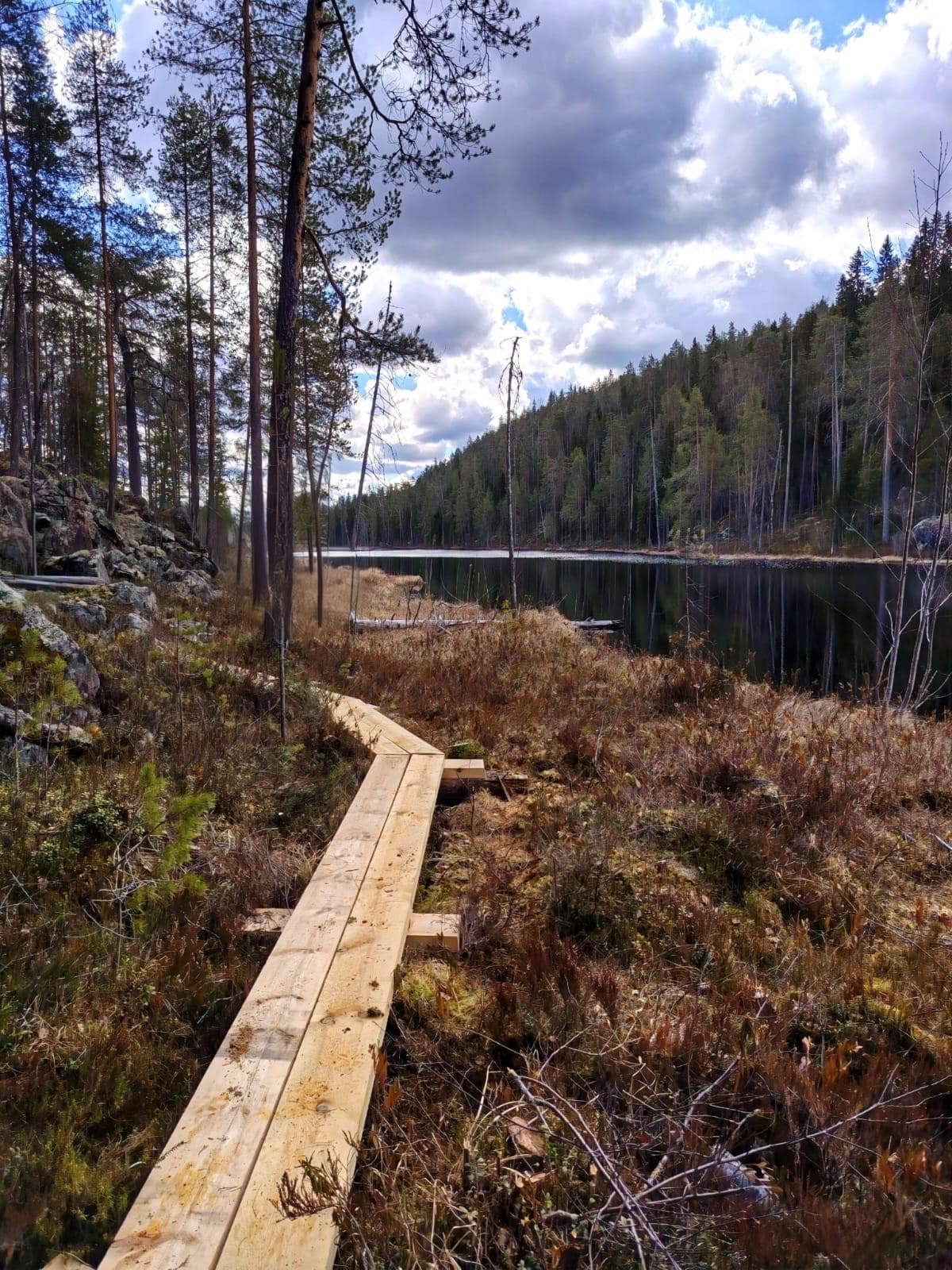 Bild: Hannu Matilainen På vänstra sidan av bilden finns spångar, i mitten myrmark, till höger en å och högst upp himmel och skog.pitkospuita, keskallä suota, oikealla joki yläreunassa taivasta ja metsää.