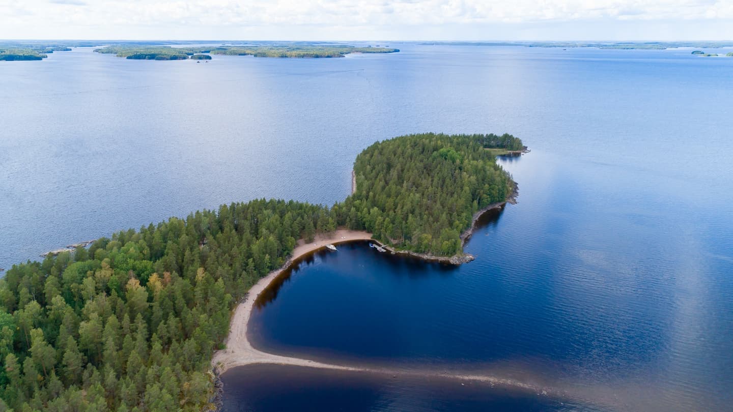 Kuva: Etelä-Karjalan virkistysaluesäätiö Ilmakuvassa on järvenselällä saaria. Lähimmässä on hiekkaranta, jossa on veneitä ja laituri.