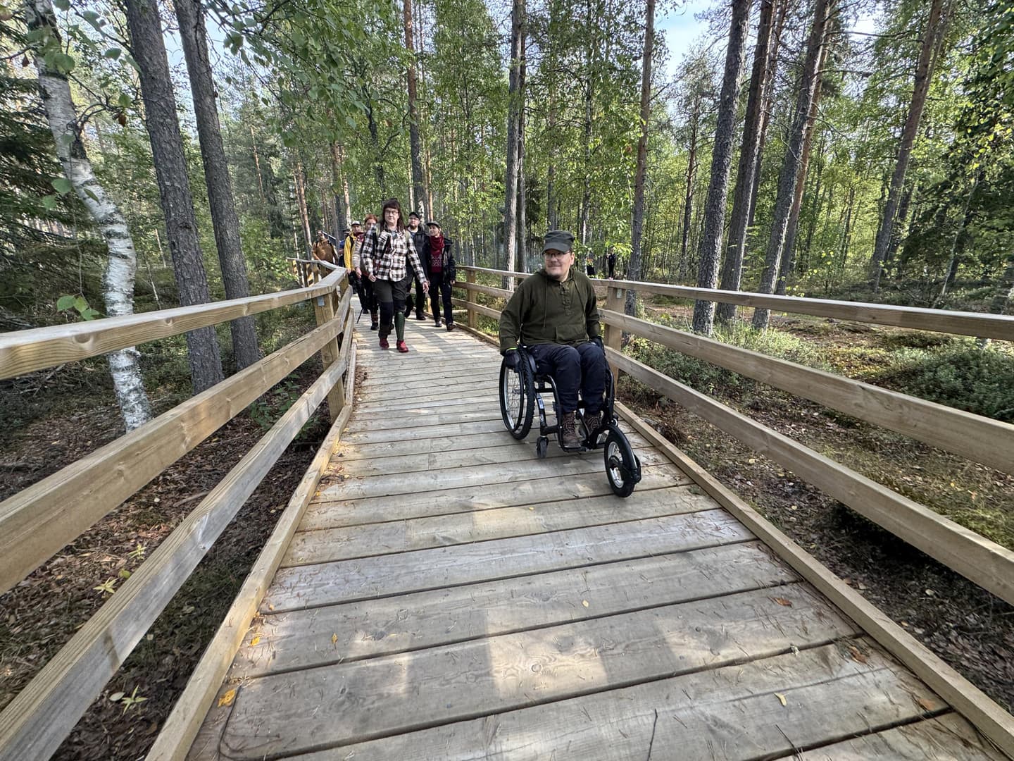 Kuva: Ilkka Numminen Ryhmä retkeilijöitä kulkee metsään rakennetulla puupolulla. Yksi kulkee pyörätuolilla.