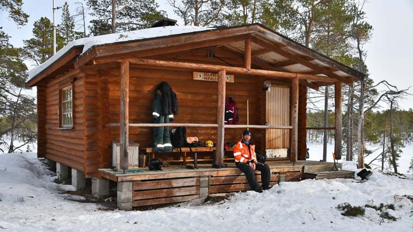 Image: Tuija Kangasniemi A hiker is sitting on the steps of the hut's porch. There is snow around the hut.