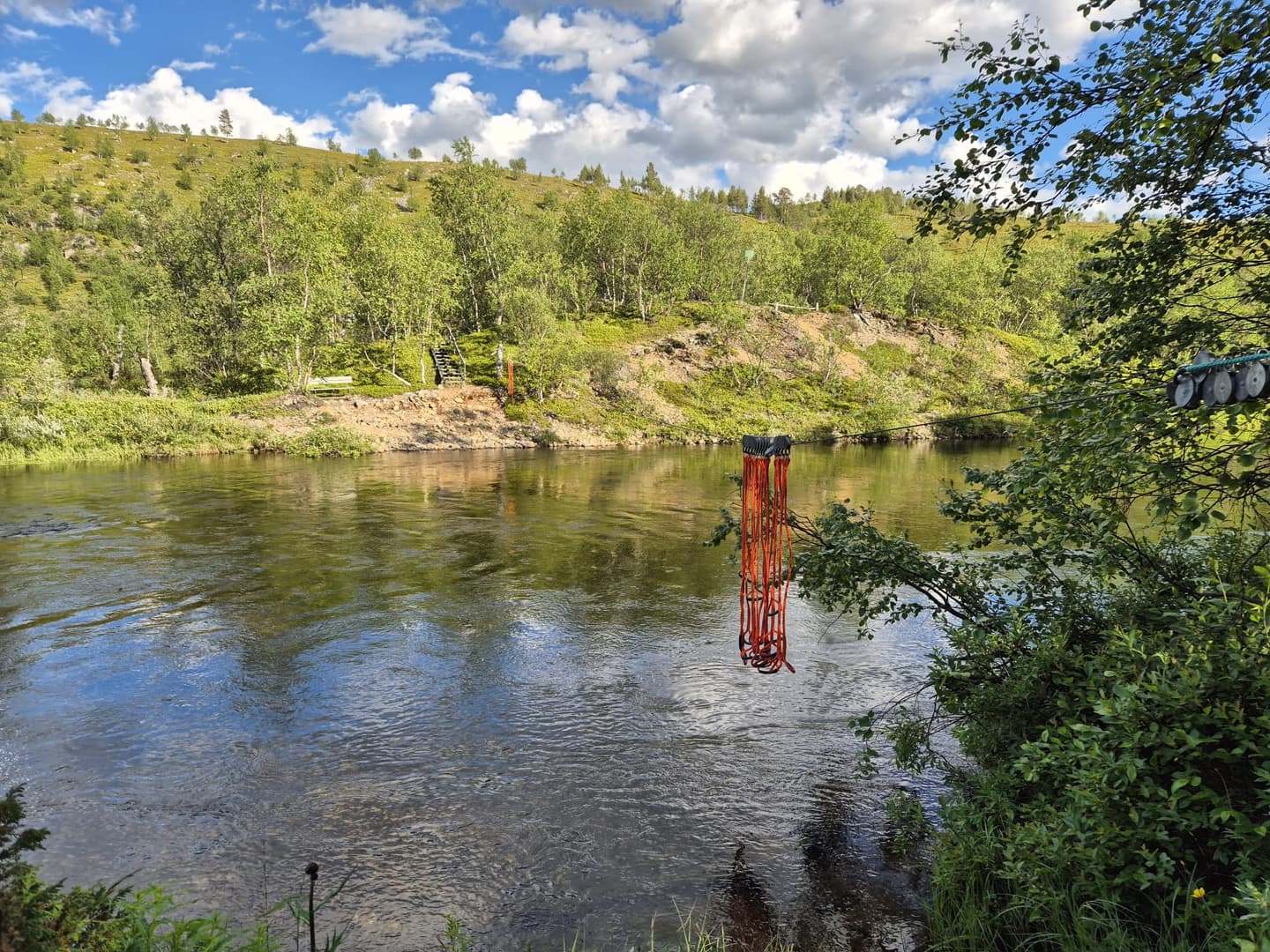 Kuva: Johanna Kulmala Joen yli on vedetty vaijeri, josta roikkuu oransseja metrinmittaisia naruja.