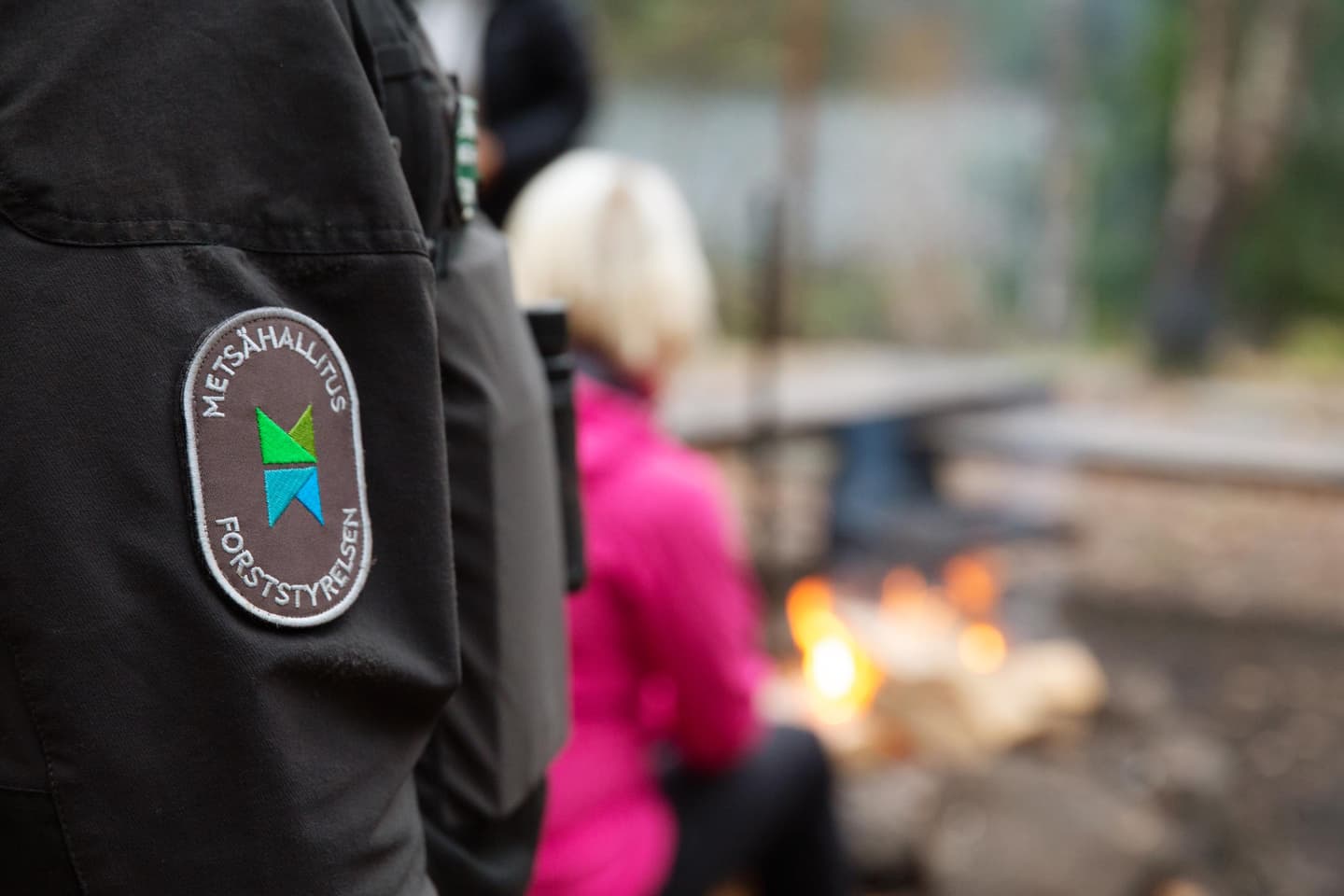 Kuva: Katri Lehtola / Keksi Metsähallituksen tunnus erätarkastajan hihamerkissä. Taustalla retkeilijä tulentekopaikalla.