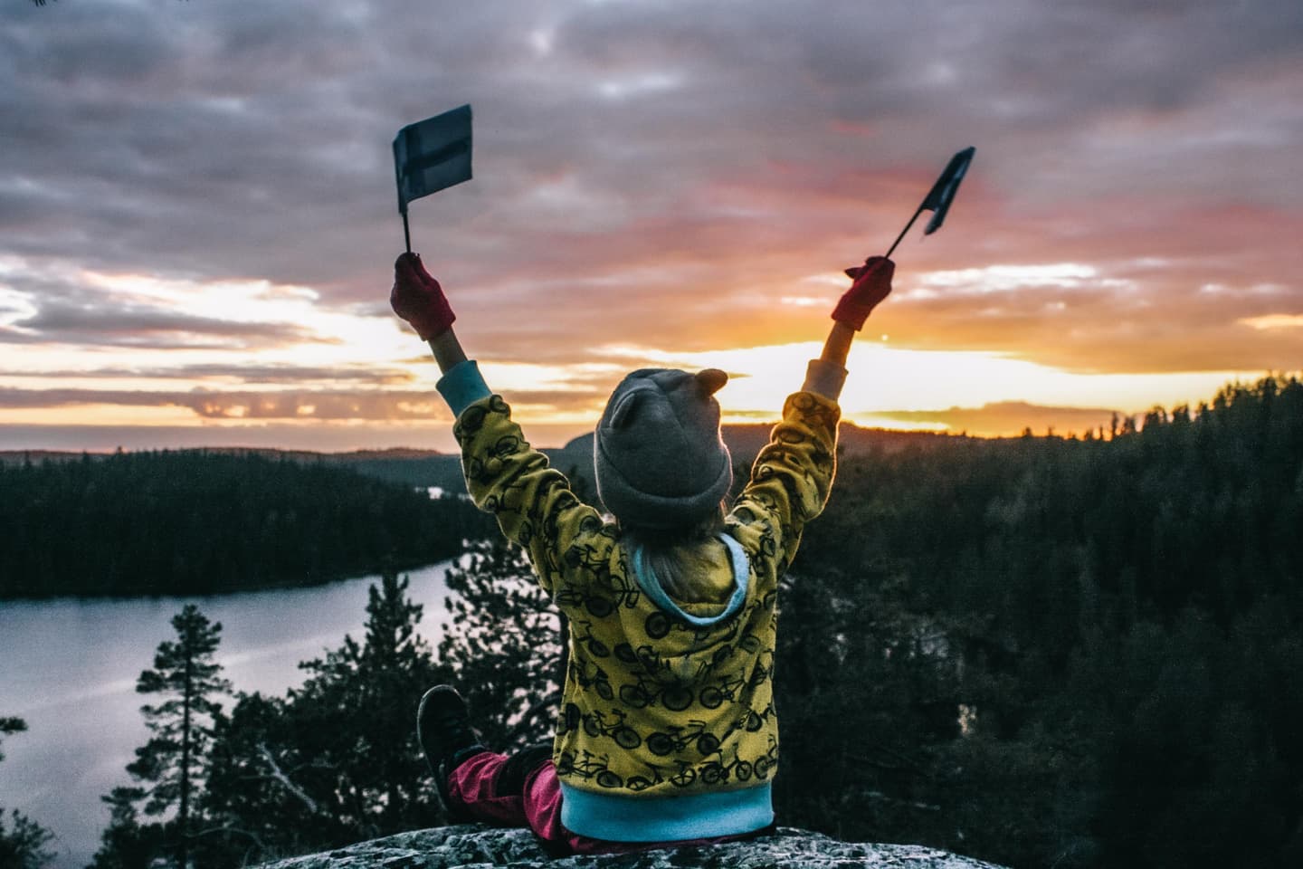 Kuva: Marjo Koramo Kuvassa etualalla kalliolla istuva lapsi pienet Suomen liput käsissään. Lapsi katsoo kamerasta pois päin auringonlaskua. Taustalla järvimaisema ja metsää.