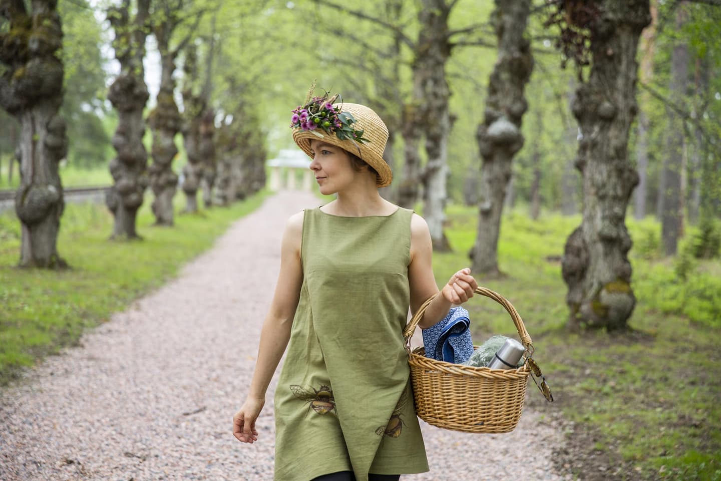 Kuva: Saara Lavi Kukkahattuinen henkilö kulkee piknikkorin kanssa puiden reunustamaa hiekkatietä pitkin.