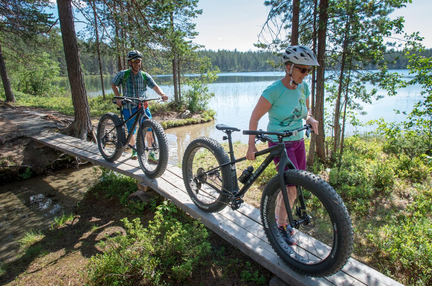 Kuva: Tuuli Turunen Kaksi retkeilijää taluttaa polkupyöriä sillan yli. Taustalla kesäinen tyyni järvimaisema.