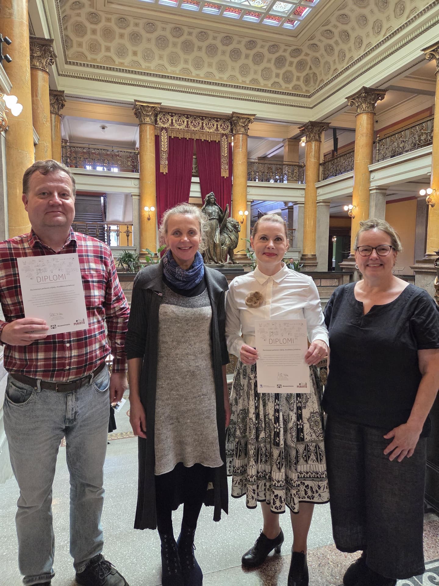 Kuva: Outileena Uotila Neljä henkilöä seisoo juhlavassa tilassa ja katsoo kohti kameraa. Kahdella on käsissään diplomi.