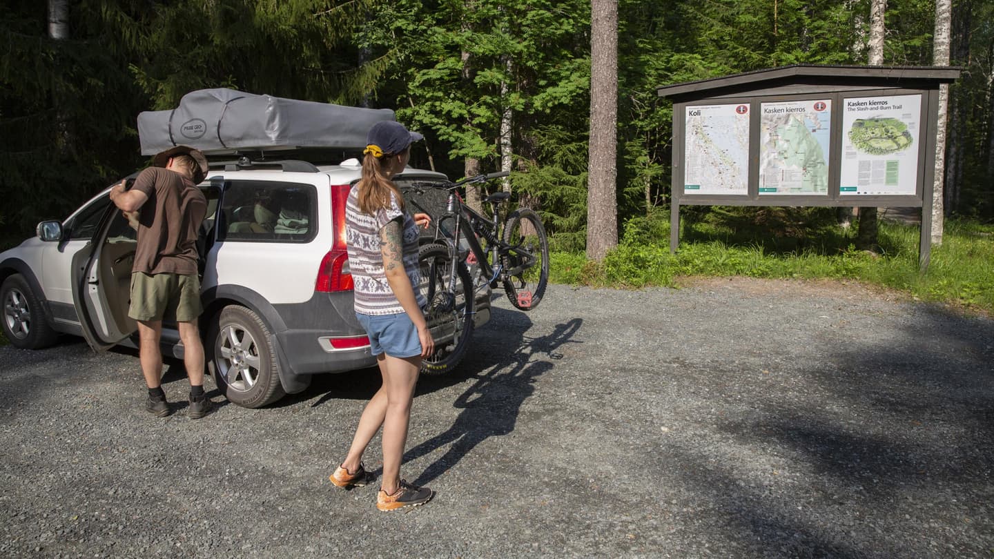 Image: Saara Lavi Two people at the Kasken kierros -Trail parking area in Koli National Park. A large three-panel information board stands at the edge of the parking area. The car shown in the photo has an unopened roof tent mounted on its roof.