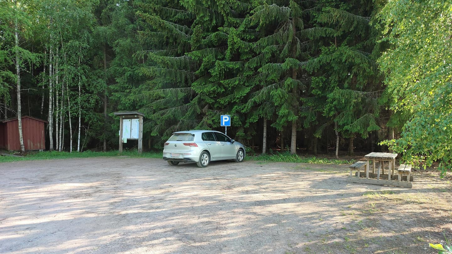 Image: Jussi Helimäki A parking area on the edge of a spruce forest, with a picnic table, information boards and a small red wooden building.
