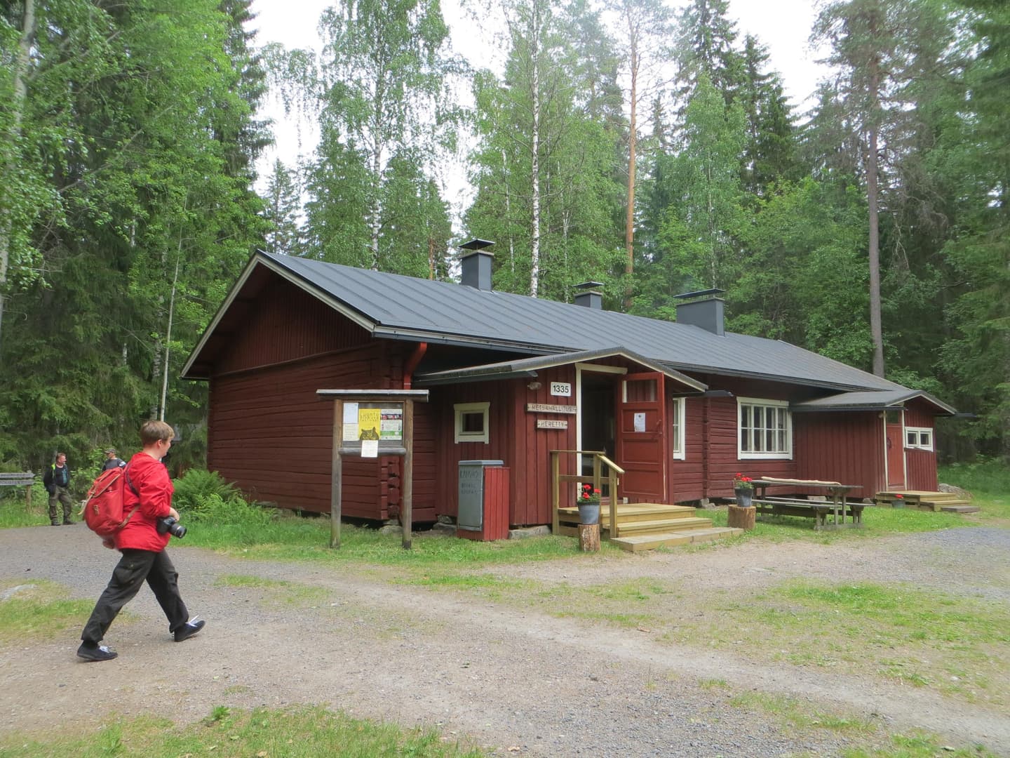Kuva: Maija Mikkola Iso hirsinen kämppärakennus kahdella oviaukolla. Kesäisessä maisemassa etualalla retkeilijä.