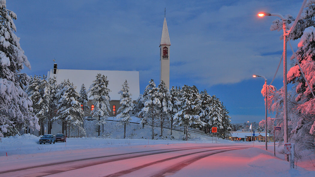Sinisen hämärä hetki kirkonkylällä. Maisema on luminen ja kuvassa on keskellä valaistu kirkko, joka on lumisten mäntyjen ympäröimä.