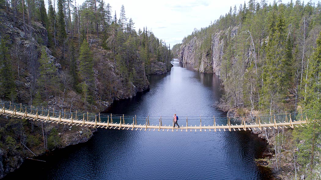 En vandrare går över en hängbro vid en ravinsjö. Bron ligger cirka 10-15 meter över vattnet. Steniga höjder reser sig vid strandkanterna.