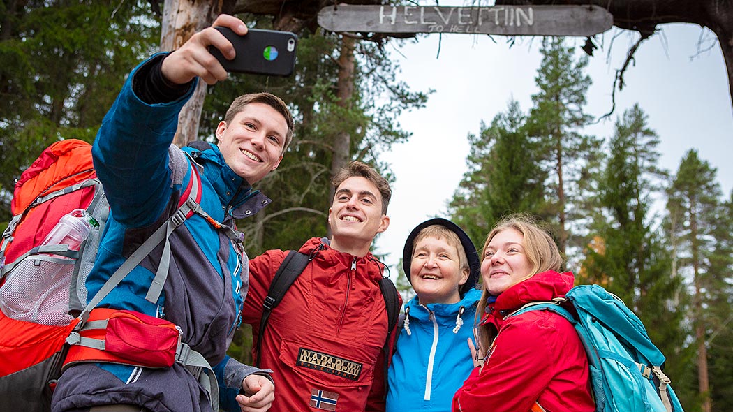 Eri-ikäisiä henkilöitä ottamassa selfie-valokuvaa. Taustalla puun rungoista rakennettu portti.