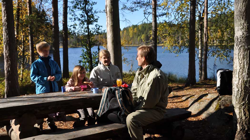 Perhe pöytäryhmän ääressä syksyisessä metsämaisemassa järven rannalla.