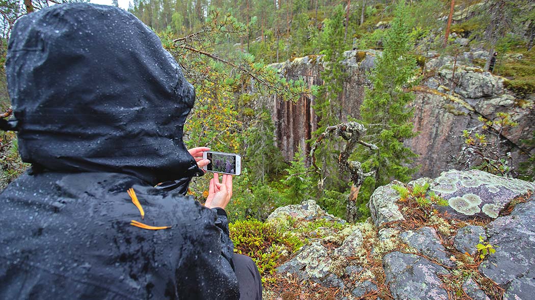 En vandrare tar en bild av en klippväggig klyfta med en mobiltelefon i regnigt väder. I förgrunden lavartade stenar och ett knotigt träd.