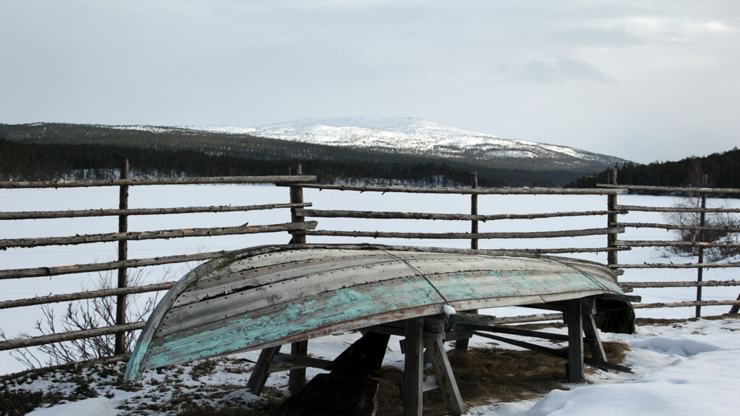 I ett vintrigt landskap ligger en båt upp och ned. I bakgrunden skymtar en snötäckt fjälltopp.