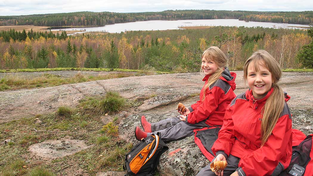 Kaksi hymyilevää lasta istuu kalliolla pullat kädessä. Taustalla aukeaa syksyinen metsä- ja järvimaisema.