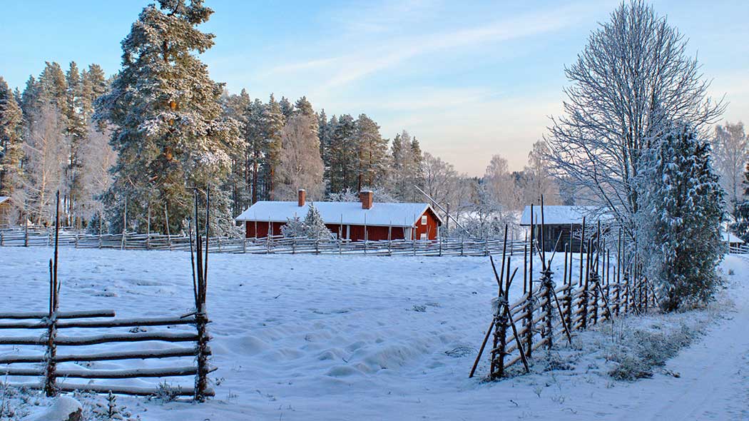 Talvinen maisema, lunta maassa, taustalla perinnetilan pihapiiri ja punainen vanha rakennus.