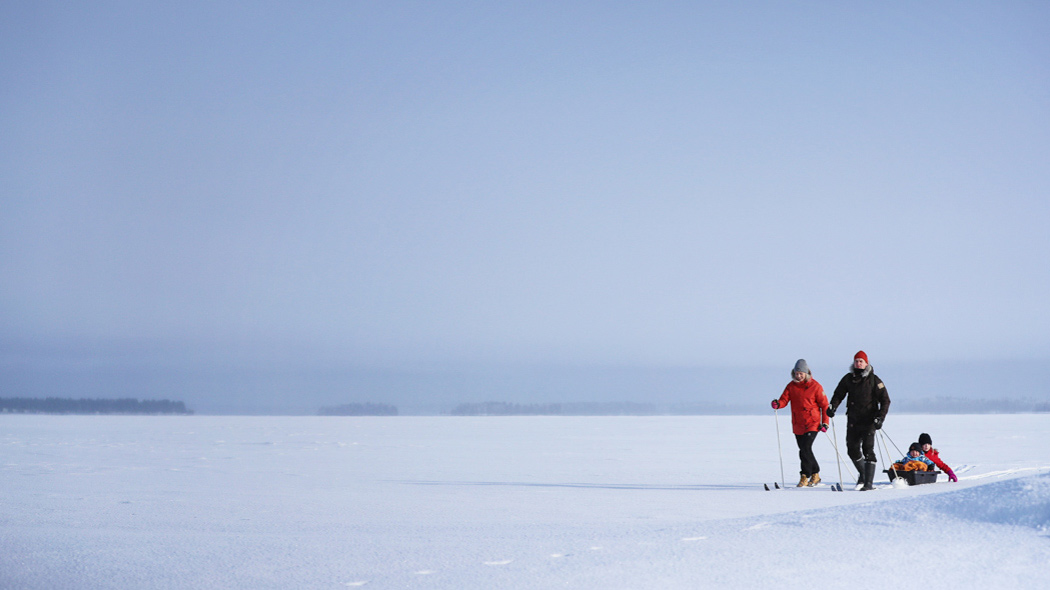 Två vuxna åker skidor på en snöig och öppen sjö. Den andra följs av en spjälsäng med två barn.