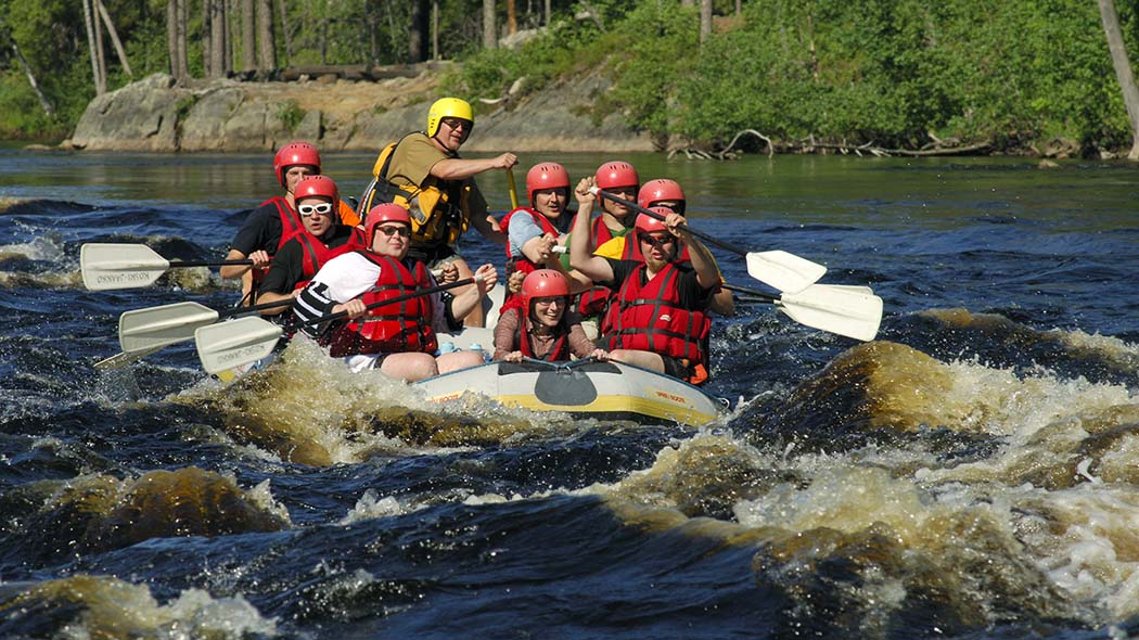 People in a rubber boat in a white water rapid. A forested rocky shore can be seen in the background.