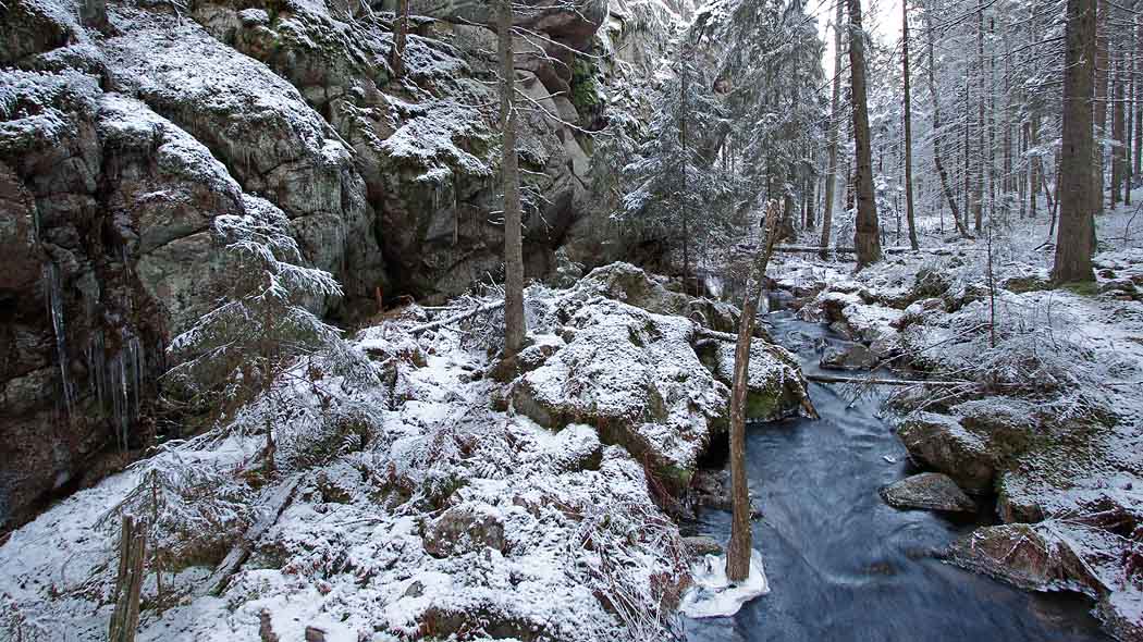 Metsässä virtaa puro, jonka vierellä on kallio. Maassa on ohut lumipeite.