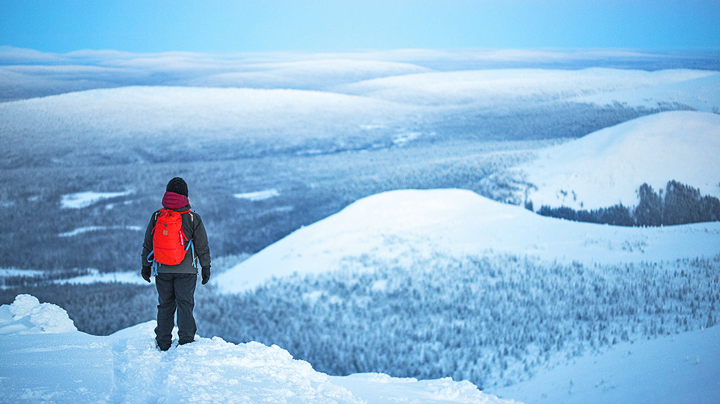 Nainen punainen reppu selässä lumisella tunturinlaella selin. Nainen katsoo horisonttiin, jossa näkyy lisää tuntureita. Tuntureilla on lunta.