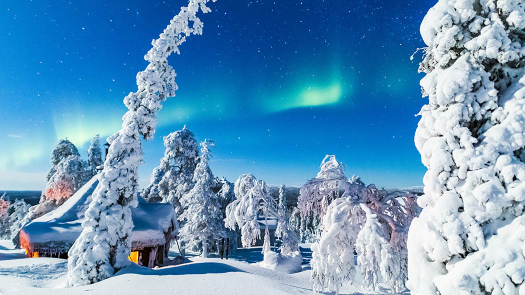 Träd täckta i ett tjockt lager snö runt en fjällkåta. Stjärnor och norrsken kan ses på natthimlen.
