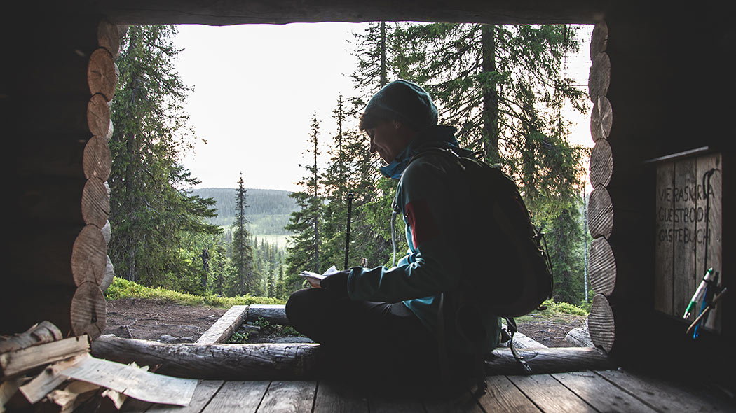 En vandrare sitter i en skjul och läser en bok. I bakgrunden kan du se landskapet med föll och träsk.