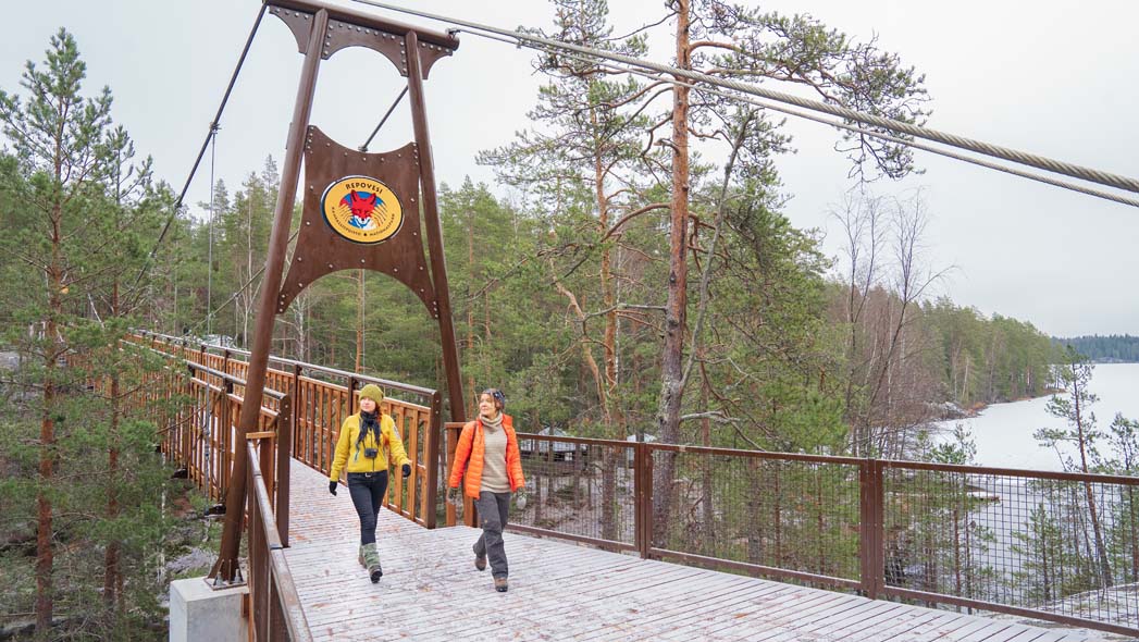 Kaksi retkeilijää astelee ohuen lumikerroksen peittämällä sillalla. Sillan päädyssä on suuri logo, jossa on ketun pää ja pihlajan lehtiä sekä teksti Repoveden kansallispuisto, Repovesi National Park.
