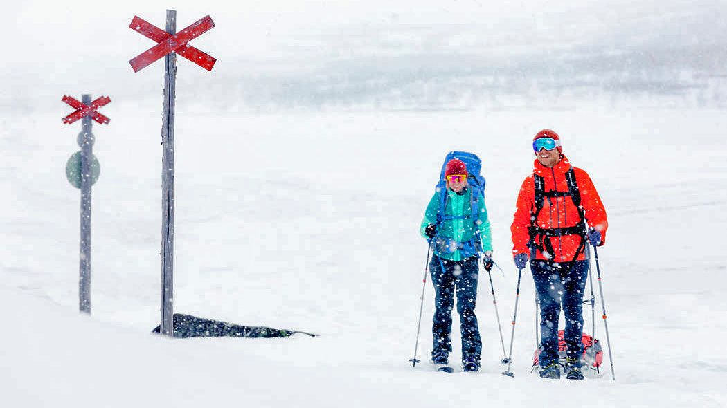 Kaksi hiihtovaeltajaa vetää ahkioita ristiviitoin merkityllä reitillä. Sataa lunta. Taustalla luminen maisema.