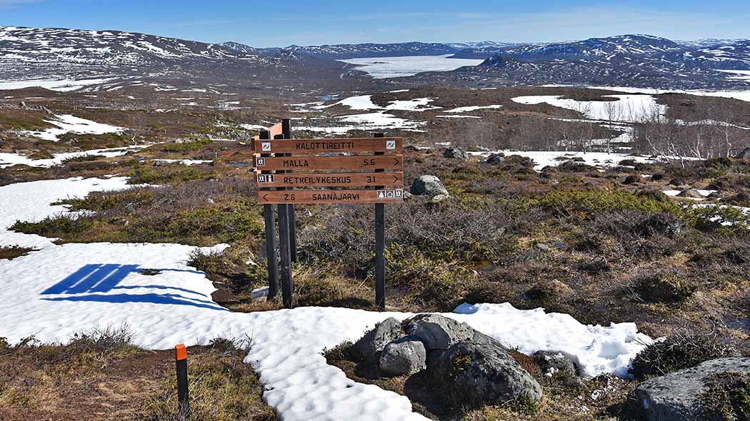 Bilden visar öppen fjällterräng, där det fortfarande ligger mycket snö. I mitten av bilden finns skyltar i trä som säger t.ex. Nordkalottleden.