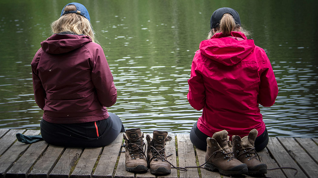 Kaksi retkeilijää istuu lankuilla veden äärellä ilman kenkiä.