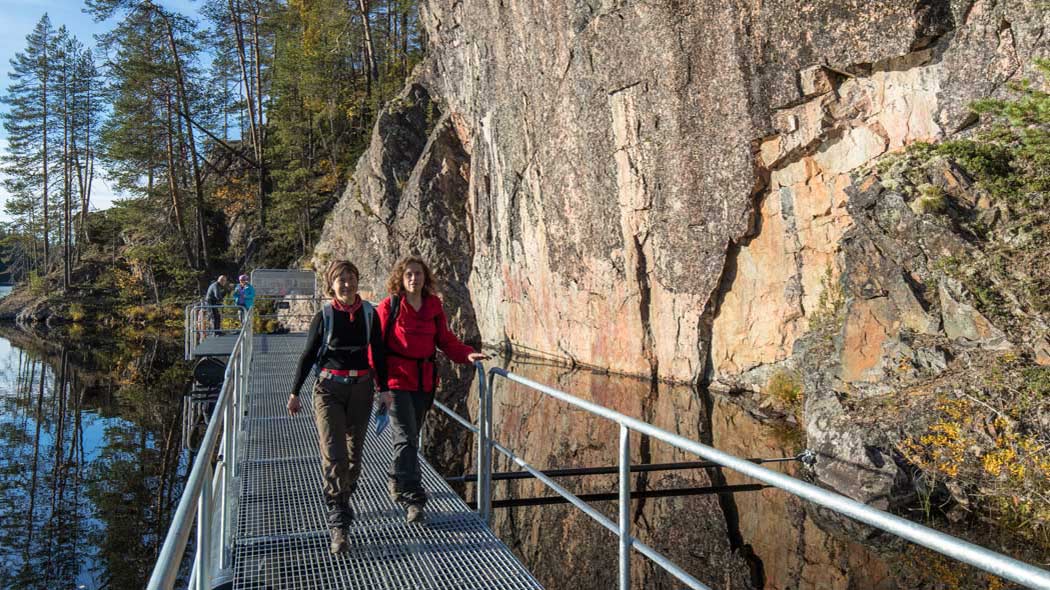 Två personer går längs en gångbro gjord av ett metallnät, gångbron följer en klippvägg vid sjöns strand. Två personer kan ses i bakgrunden.