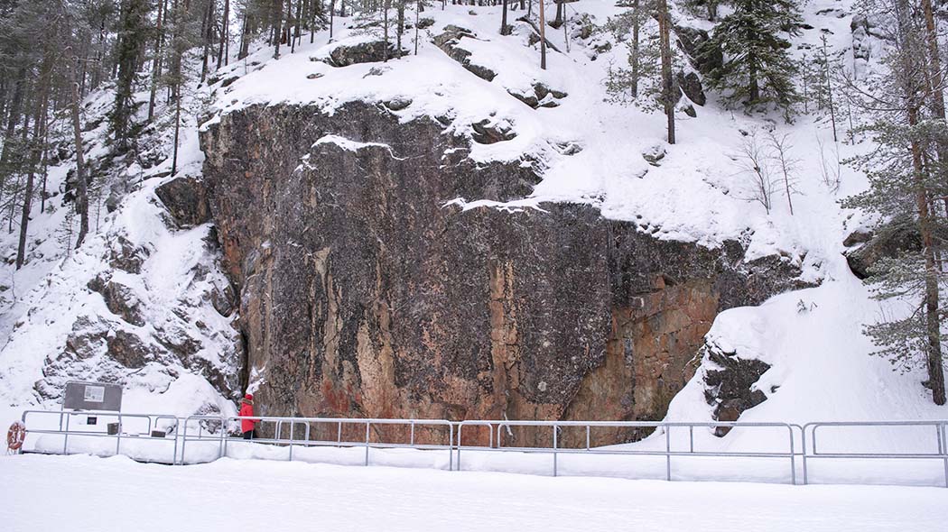 Vandraren tittar på målningarna på bergväggen. Det finns en gångbro gjord av metall vid berget. Det ligger snö på marken.