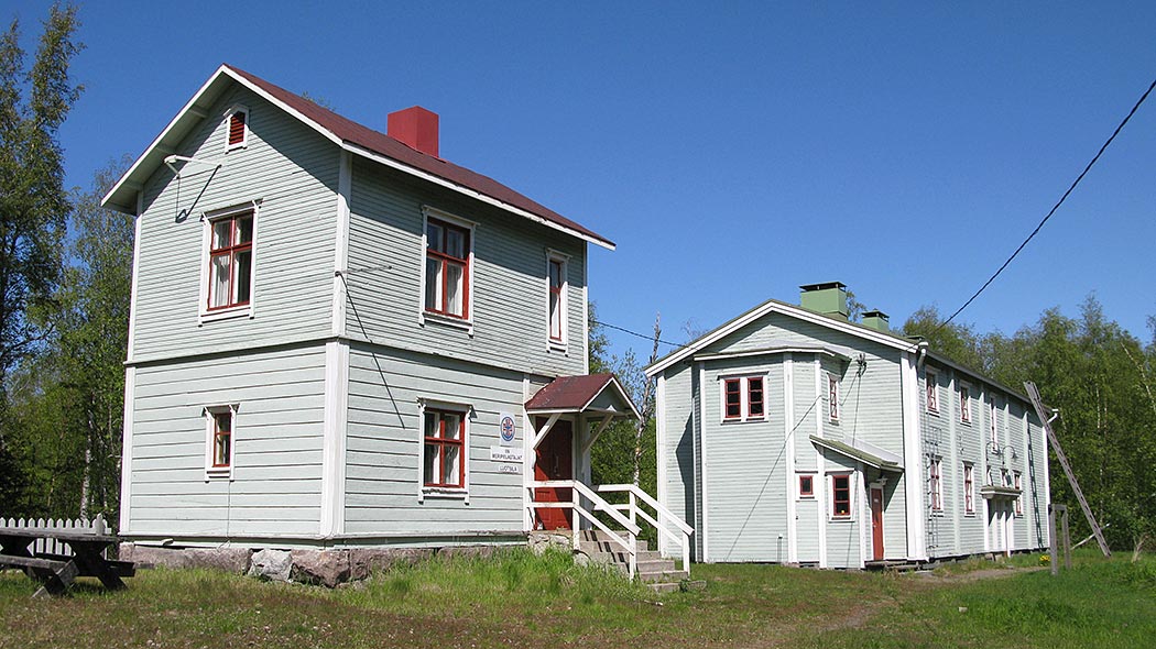 Två stycken tvåvåningsbyggnader i samma färg bredvid varandra.  Den större byggnaden har en sockel av stora stenar. Det finns en sittgrupp gjord i trä på gården.