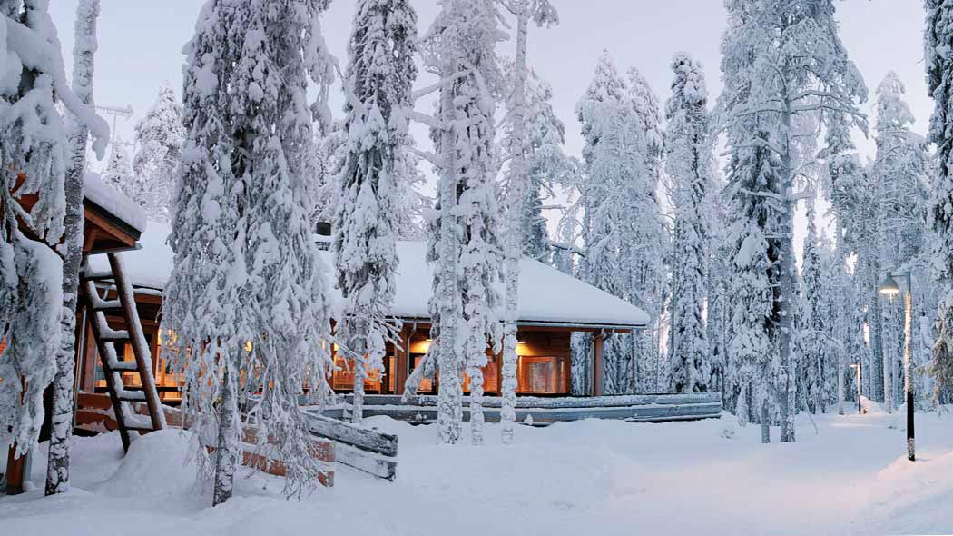 Hirsirakennus, jossa on valaistus päällä sisätiloissa, seisoo suurten puiden keskellä. Lunta on paksusti maassa ja puissa.