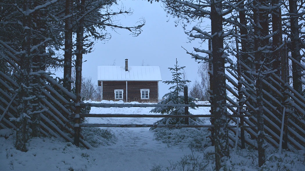 Riukuaidan veräjän takaa näkyy vaaran laella oleva talo. Maassa, puissa ja talon katolla on lunta.